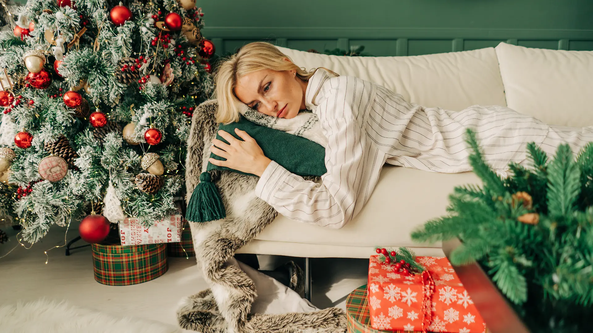 Frau liegt entrüstet und erschöpft im Schlafanzug auf ihrer Couch, die in einem geschmückten Weihnachtswohnzimmer steht