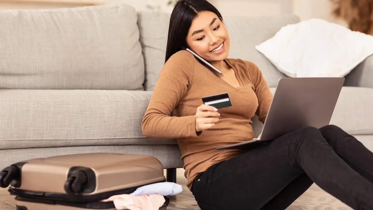 Eine junge, asiatische Frau steckt mitten in den Urlaubsvorbereitungen. Sie telefoniert am Handy und hält ihre Kreditkarte zur Buchung am Laptop bereit. Neben ihr liegt schon der Reisekoffer.