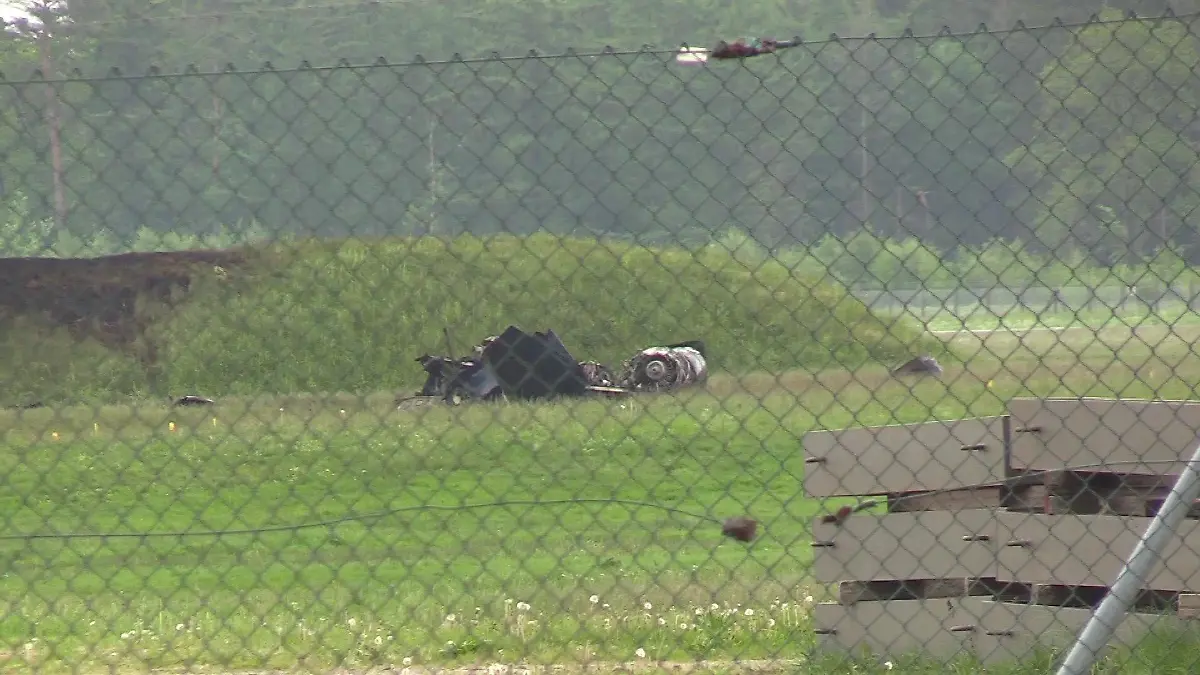 Hinter einem Drahtmaschenzaun, der den Flugplatz umrandet, liegen Trümmerteile auf einer grünen Wiese.
