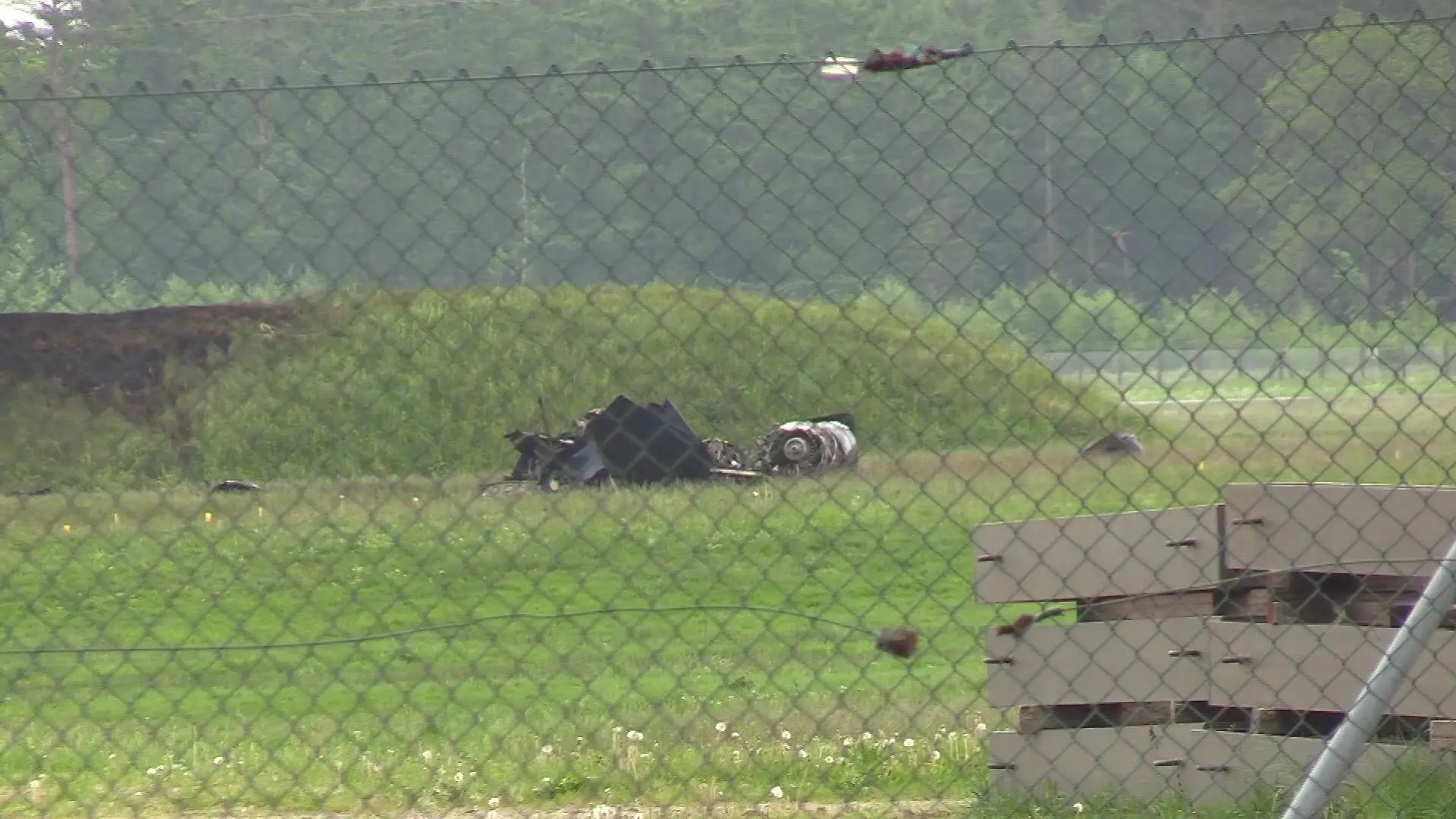 Hinter einem Drahtmaschenzaun, der den Flugplatz umrandet, liegen Trümmerteile auf einer grünen Wiese.