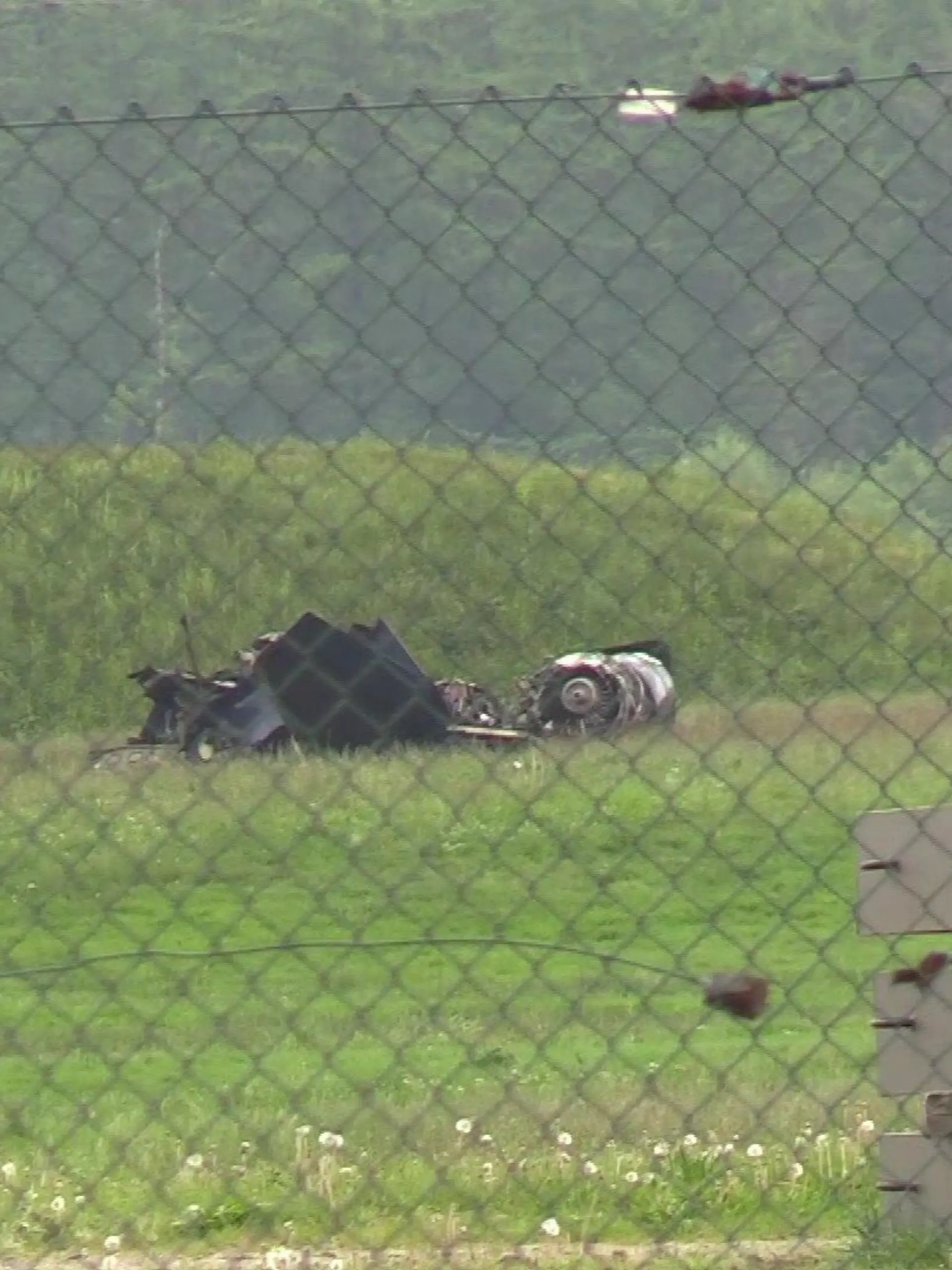 Hinter einem Drahtmaschenzaun, der den Flugplatz umrandet, liegen Trümmerteile auf einer grünen Wiese.