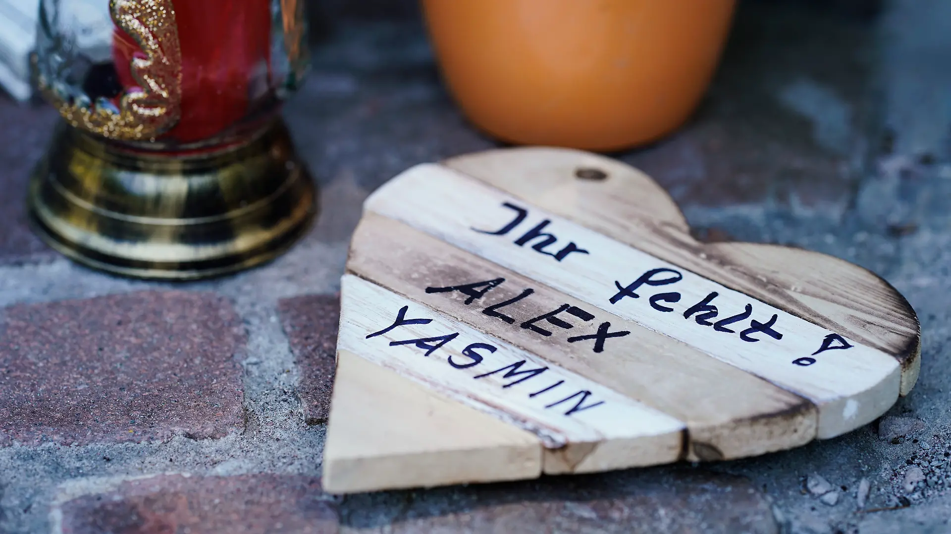 Auf einem Holzbrett in Herzform ist an einer Landstraße zwischen Kusel und Ulmet an einer Gedenkstätte für zwei getötete Polizisten die Aufschrift „Ihr fehlt! Alex Yasmin“ aufgebracht.