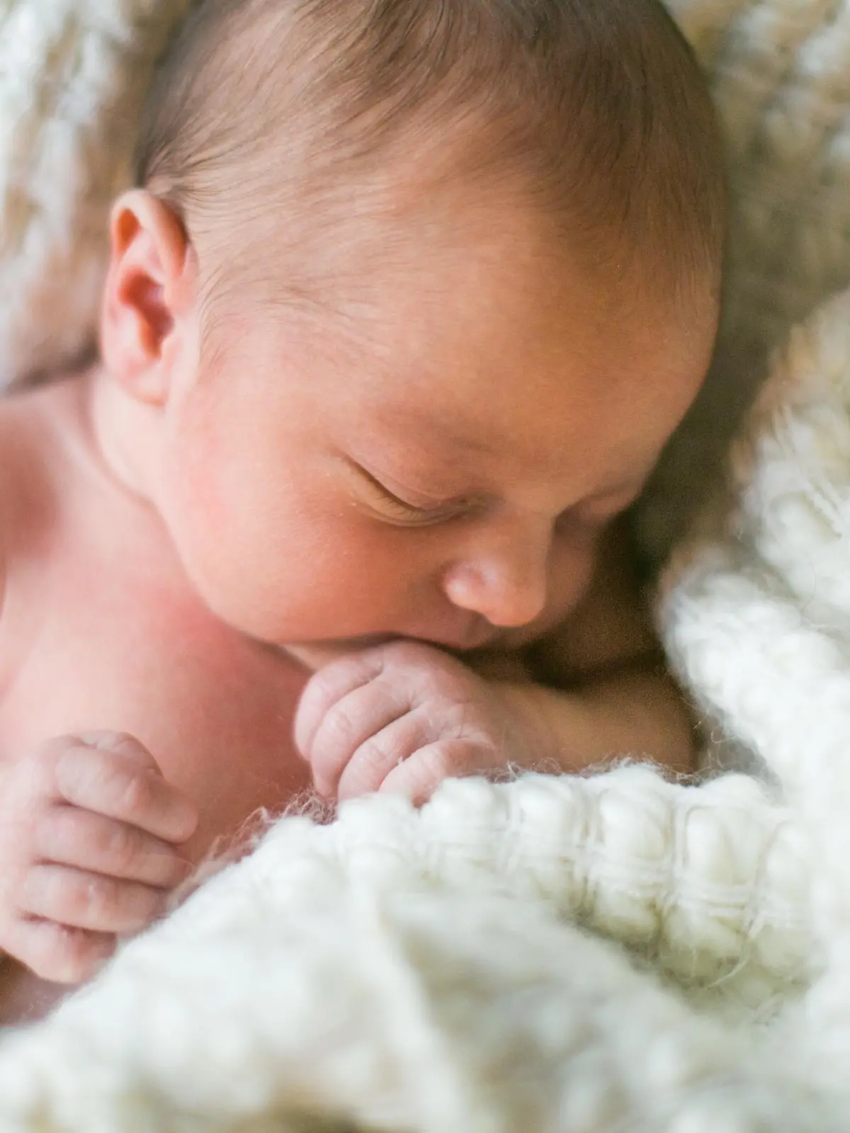 A brand new one week old baby boy sleeps wrapped in a white fuzzy blanket. He is peaceful and his hand rests near his mouth.