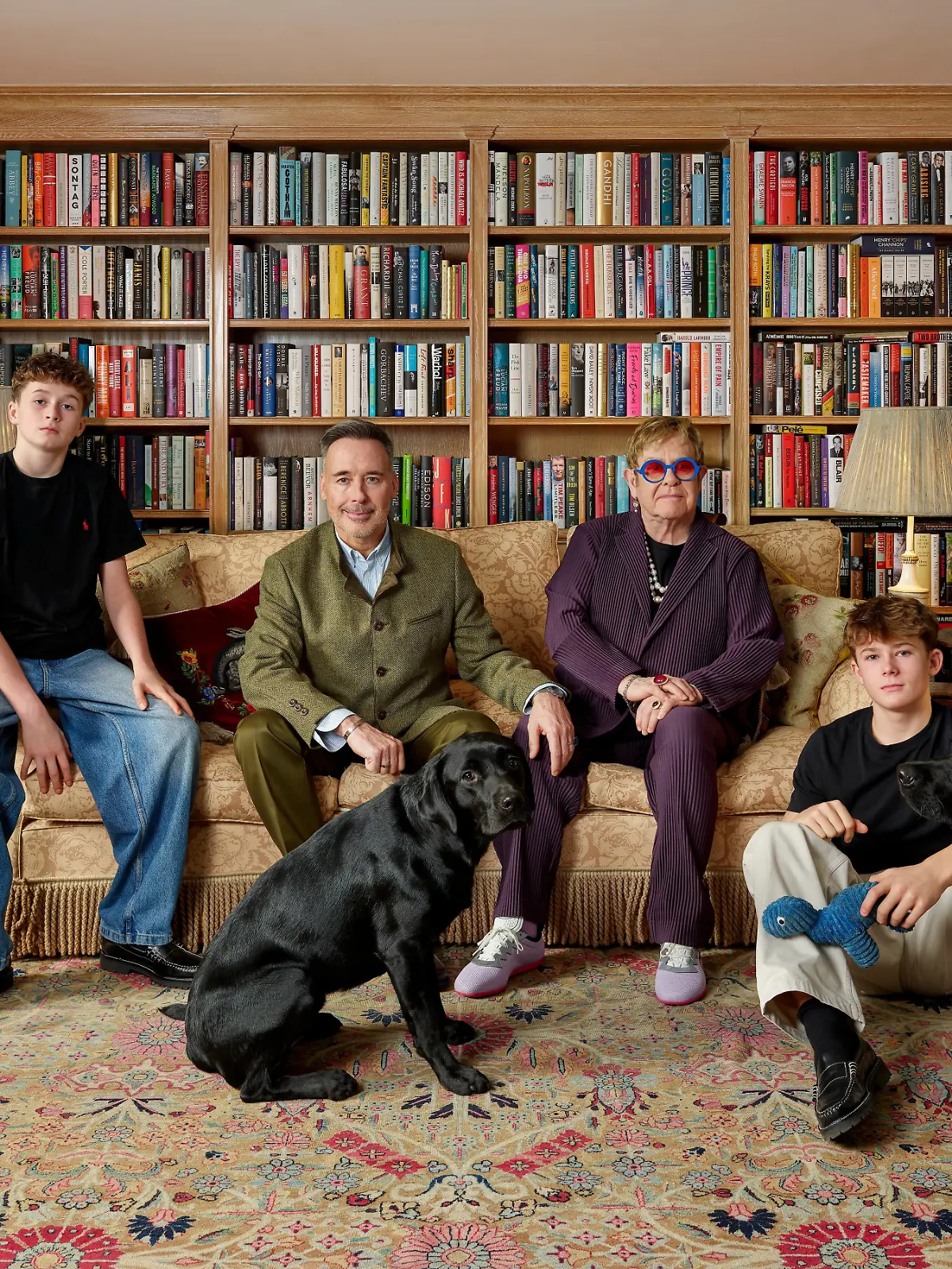 Das Bild zeigt die vierköpfige Familie beisammen auf einem Sofa inmitten von Bücherregalen. 