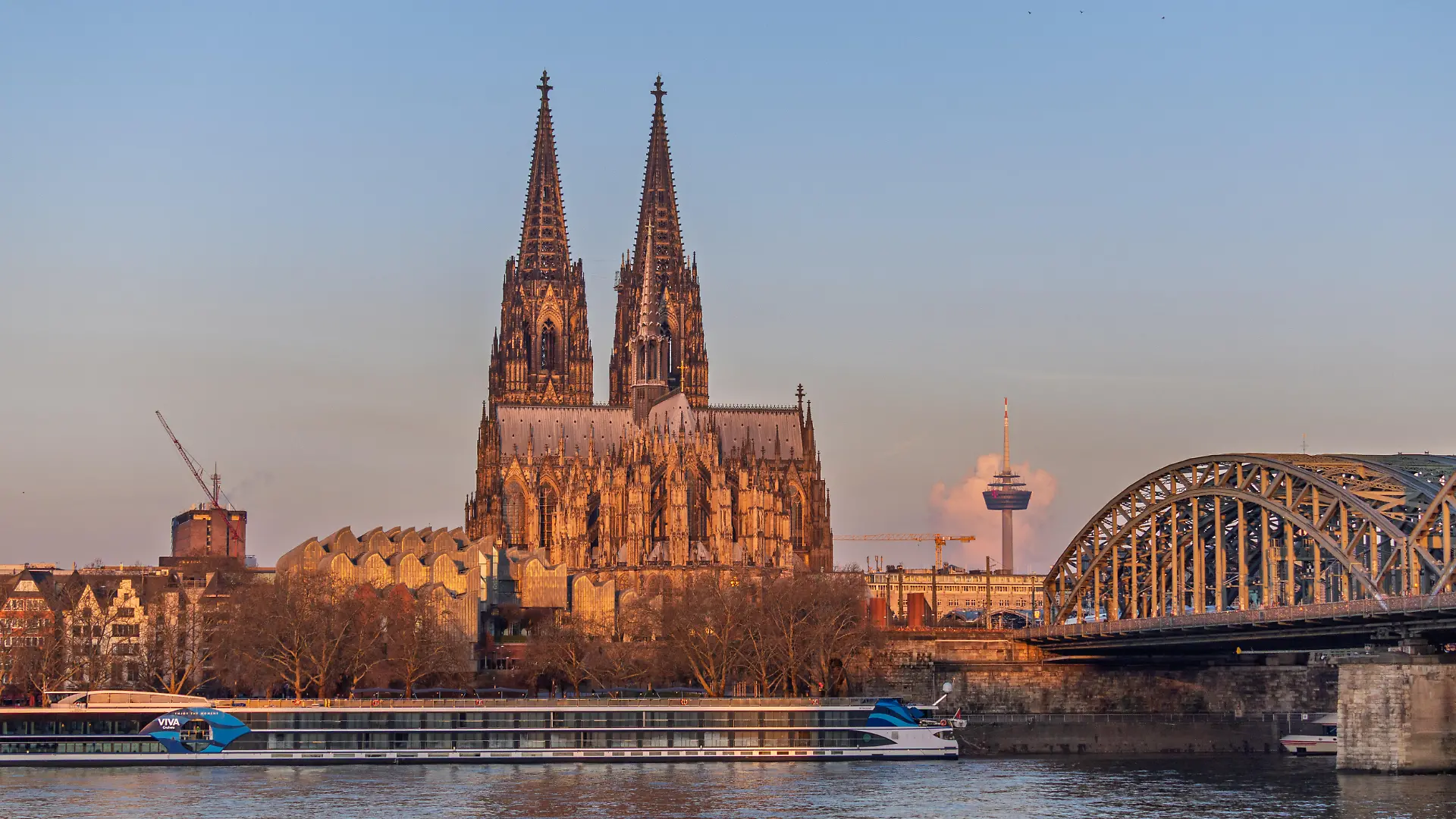 Sonnenaufgang in Köln: Blick vom Deutzer Ufer über den Rhein auf den Kölner Dom, die Hohenzollernbrücke und den Fernsehturm Colonius im Hintergrund. Die ersten Sonnenstrahlen tauchen die Kathedrale und die Fassade des Museum Ludwig in warmes Morgenlicht. Am Rheinufer liegt ein Flusskreuzfahrtschiff vor Anker.