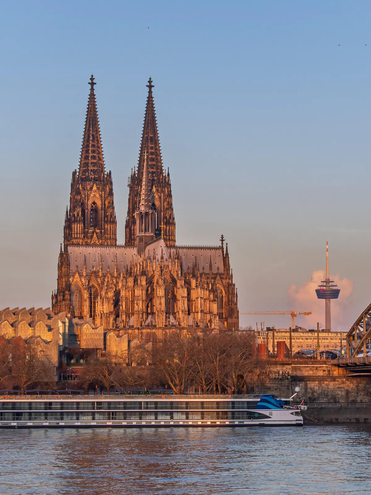 Sonnenaufgang in Köln: Blick vom Deutzer Ufer über den Rhein auf den Kölner Dom, die Hohenzollernbrücke und den Fernsehturm Colonius im Hintergrund. Die ersten Sonnenstrahlen tauchen die Kathedrale und die Fassade des Museum Ludwig in warmes Morgenlicht. Am Rheinufer liegt ein Flusskreuzfahrtschiff vor Anker.