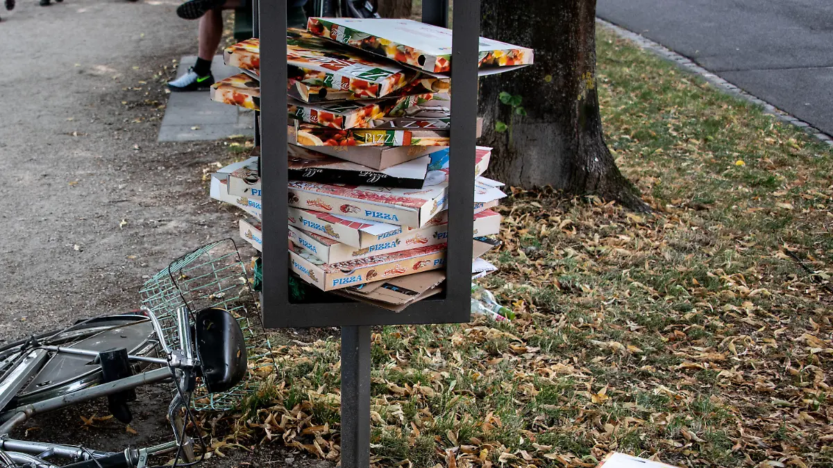 25.07.2020, Bayern, Regensburg: Zahlreiche Pizzakartons sind in einem Stahlgestänge für einen Mülleimer in einer Parkanlage gestapelt und liegen teilweise auf dem Rasen. In städtischen Parks und Grünanlagen stapelt sich der Müll von To-go-Verpackungen. Besonders an den sperrigen Pizzakartons stören sich viele Kommunen. Kann eine Pfandregelung helfen? Foto: Matthias Balk/dpa +++ dpa-Bildfunk +++