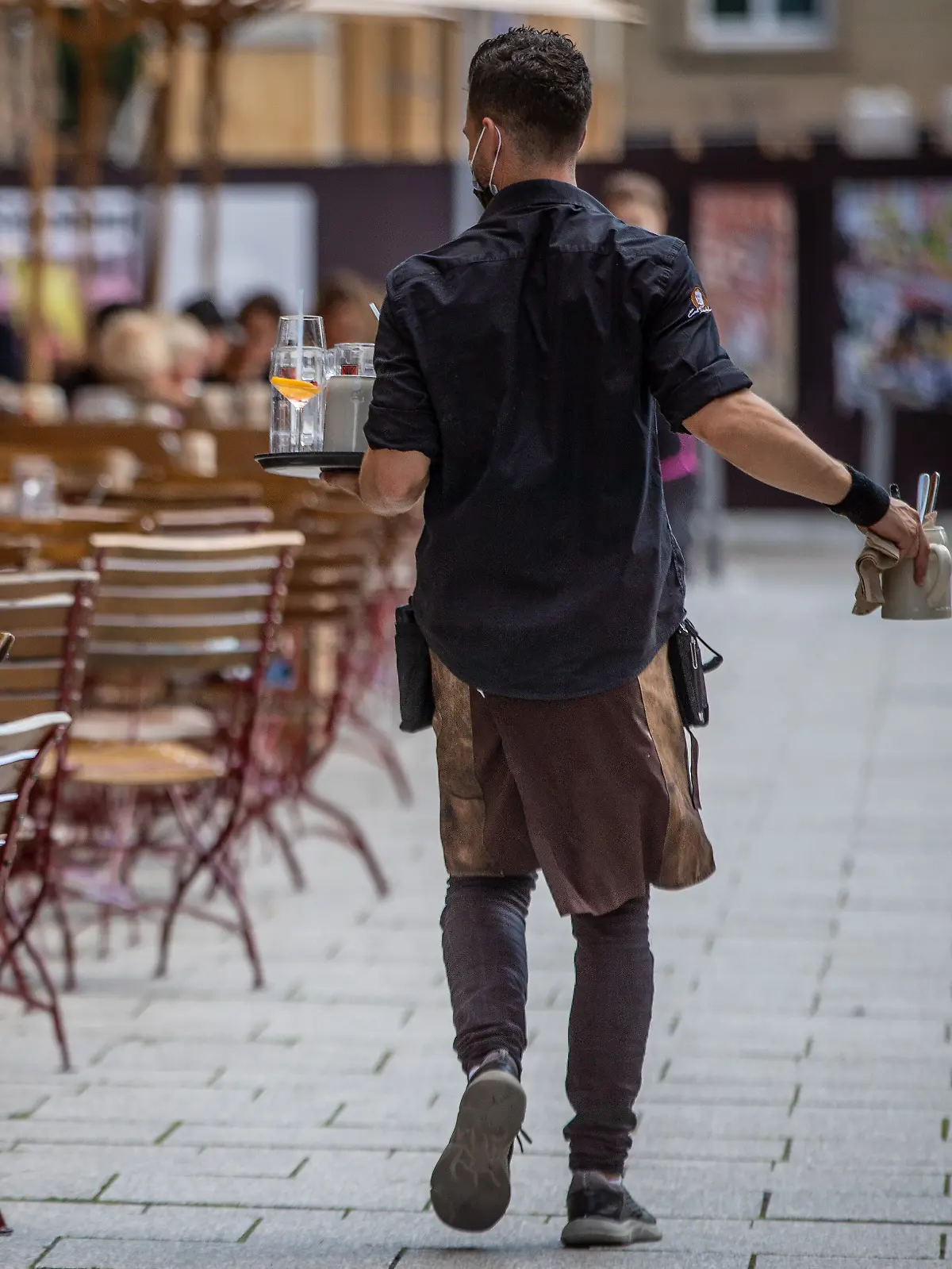Eine Servicekraft läuft im Außenbereich eines Restaurants an Gästen vorbei. Experten im Gastgewerbe und der Landwirtschaft sehen die Ausstattung mit Saisonkräften in Sachsen-Anhalt in den jeweiligen Branchen verbessert. (zu dpa: «Verbände sehen Situation mit Saisonkräften verbessert») +++ dpa-Bildfunk +++