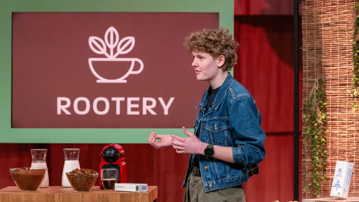 Die Höhle der Löwen: Junge Frau mit kurzen Locken und Jeansjacke steht vor dem Kaffeelogo „Rootery” und erklärt ihr Konzept