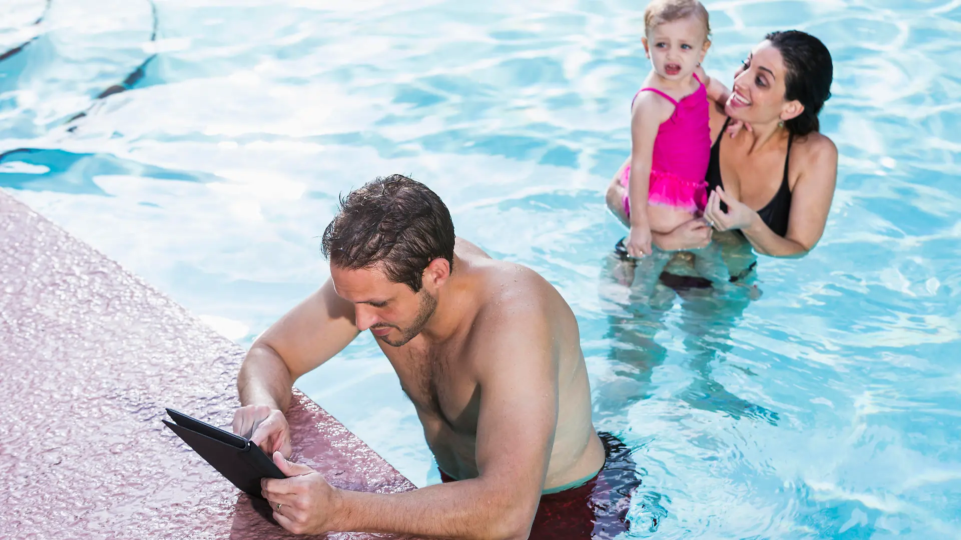 Ein Mann hängt im Schwimmbad am Beckenrand und schaut auf sein iPad, im Hintergrund sieht man eine Frau mit Kind.