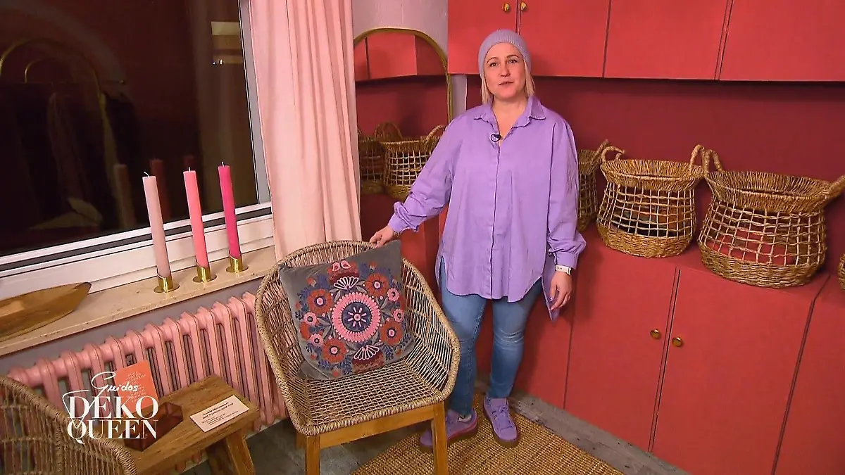 Guidos Deko Queen: Junge Frau in Jeans, lilafarbener Bluse und Mütze steht vor einem roten Schrank neben einem Korbsessel