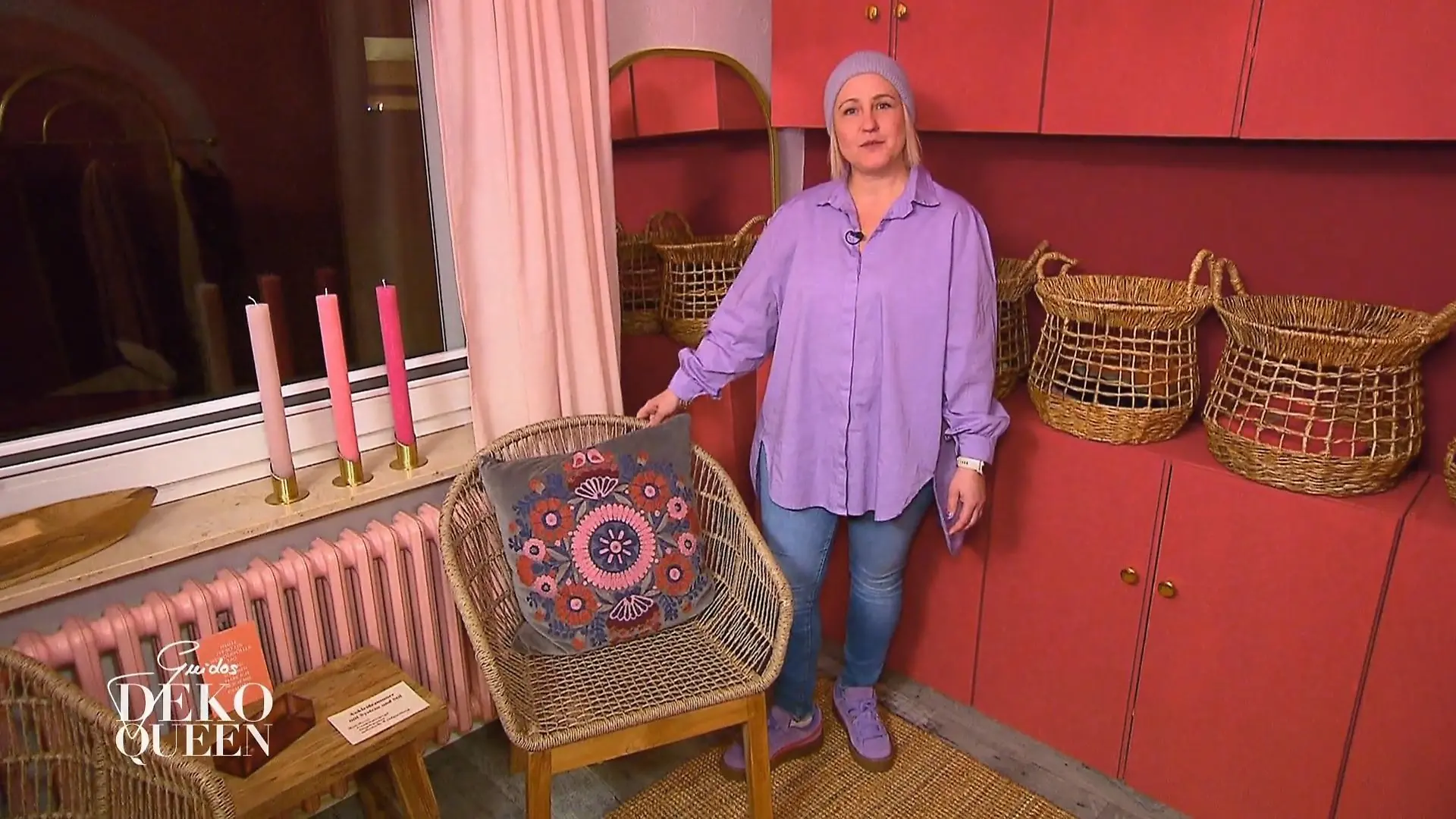 Guidos Deko Queen: Junge Frau in Jeans, lilafarbener Bluse und Mütze steht vor einem roten Schrank neben einem Korbsessel