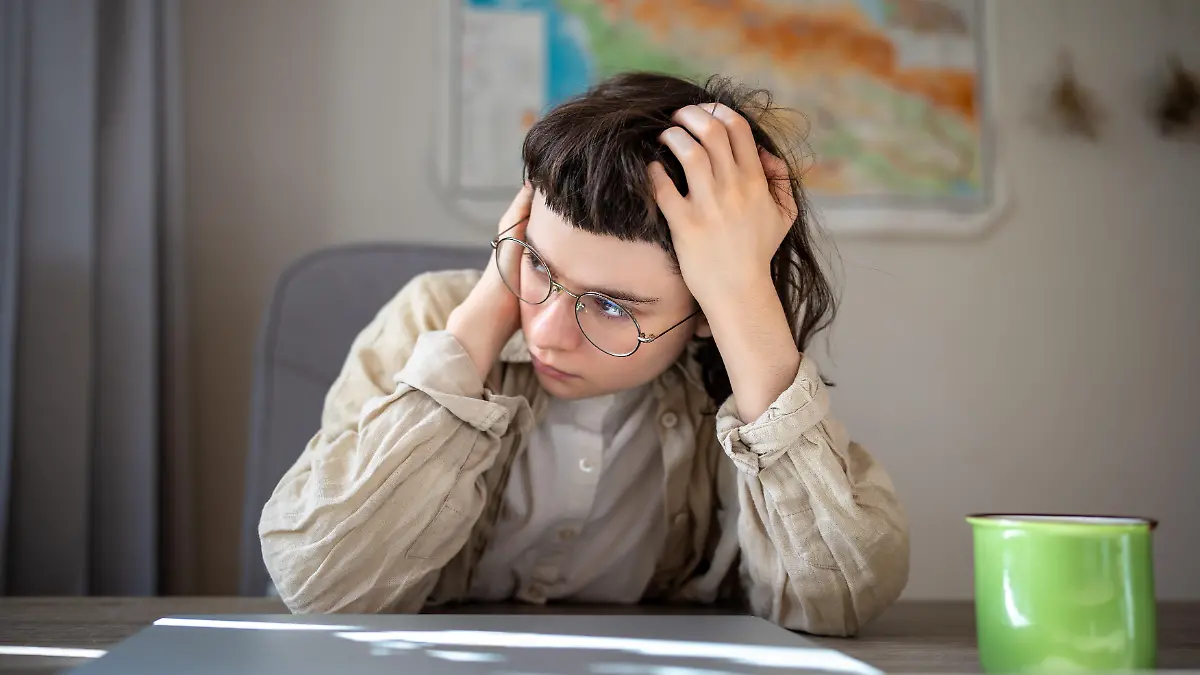 Tired teen girl in glasses high school student worried about exams sitting at desk at home. Girl holds head after sleepless night drinks coffee tries awake. Insomnia, lack of sleep, depressed mood. || Modellfreigabe vorhanden