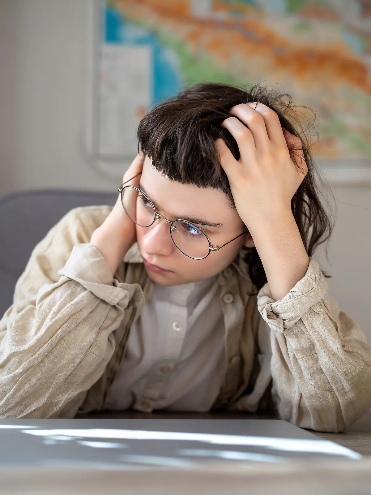 Tired teen girl in glasses high school student worried about exams sitting at desk at home. Girl holds head after sleepless night drinks coffee tries awake. Insomnia, lack of sleep, depressed mood. || Modellfreigabe vorhanden