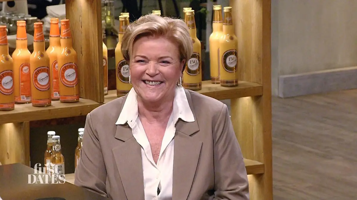 First Dates – Ein Tisch für zwei: Kandidatin Ilona - eine gepflegte lächelnde Frau über 60 mit Kurzhaarschnitt, weißer Bluse und Blazer - an der Bar