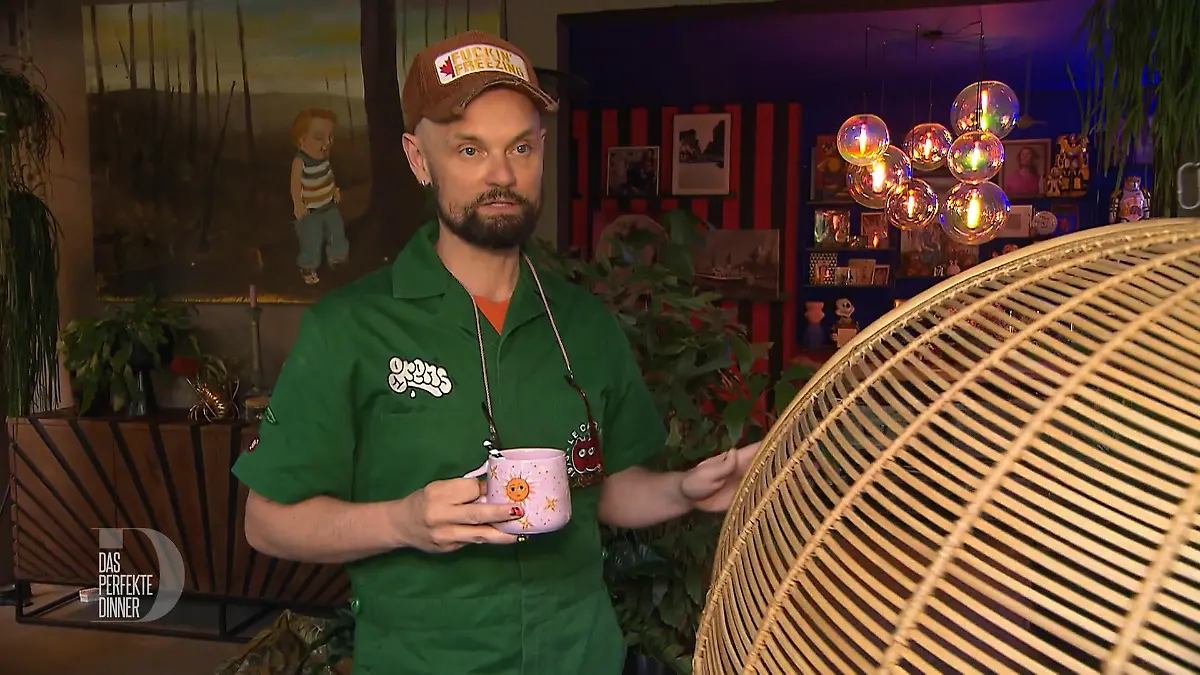 Das perfekte Dinner: Schlanker Mann mit Bart, braunem Käppi, grünem Overall und rosa Tasse in der Hand steht neben einem Korbsessel