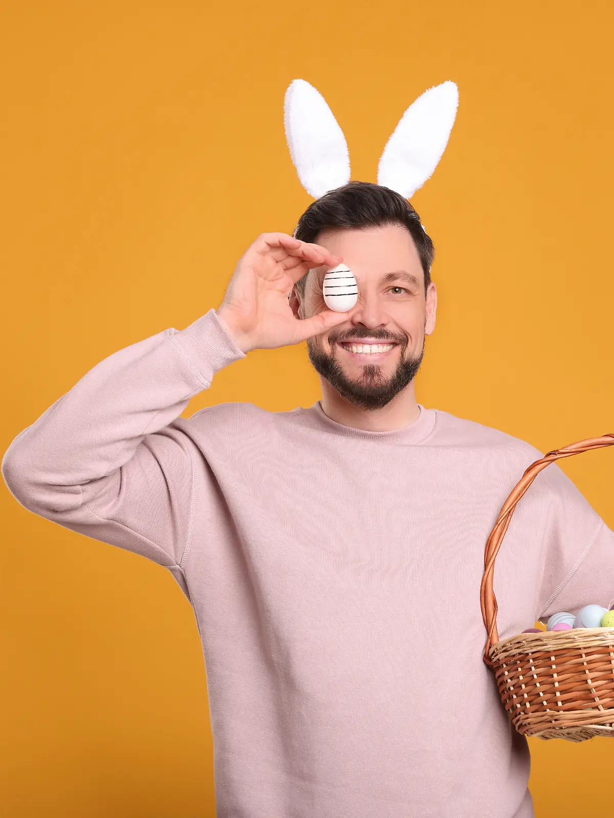 Happy man in bunny ears headband holding wicker basket with painted Easter eggs on orange background