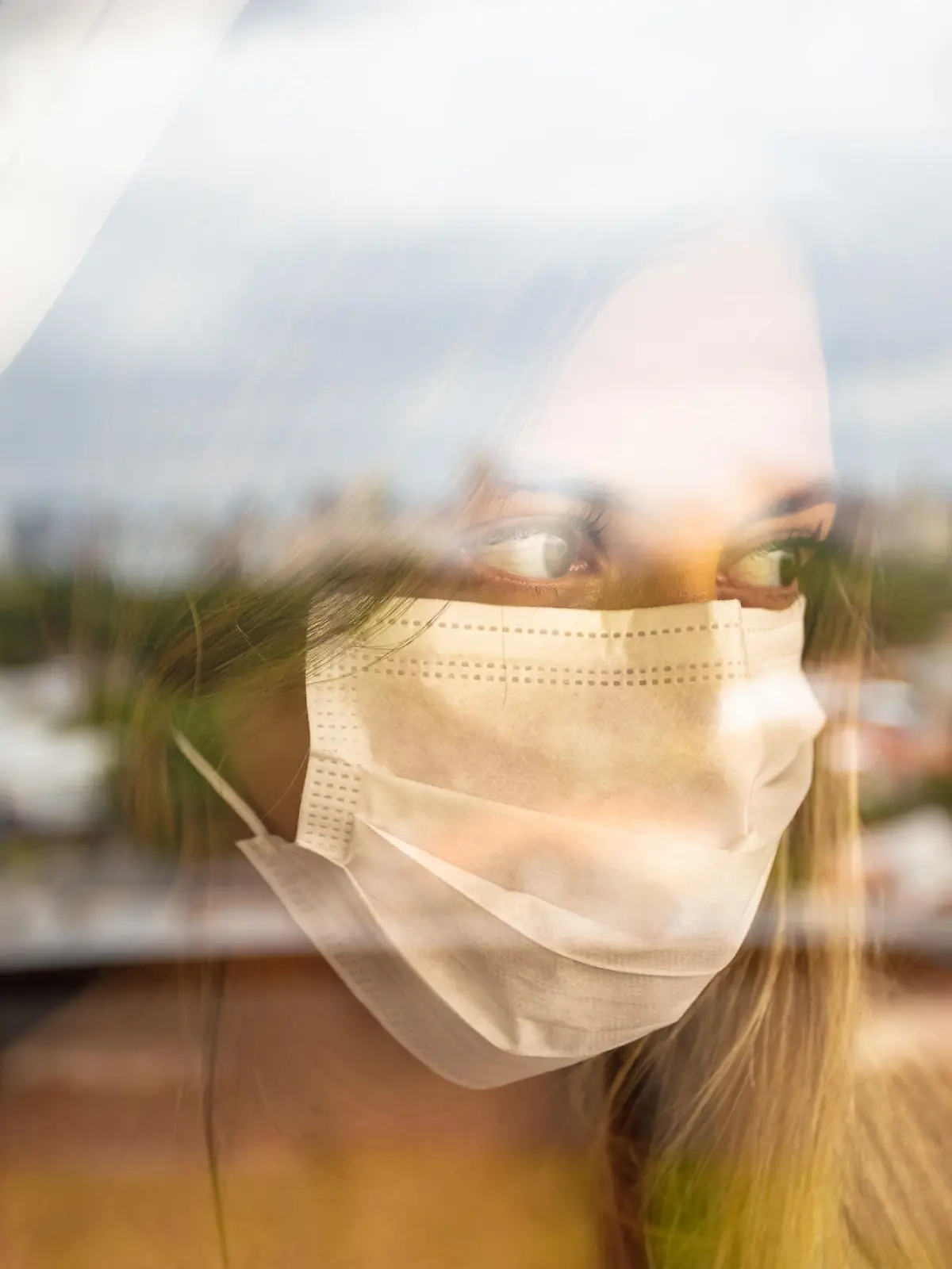 Junge Frau mit Maske steht vor ihrem Fenster