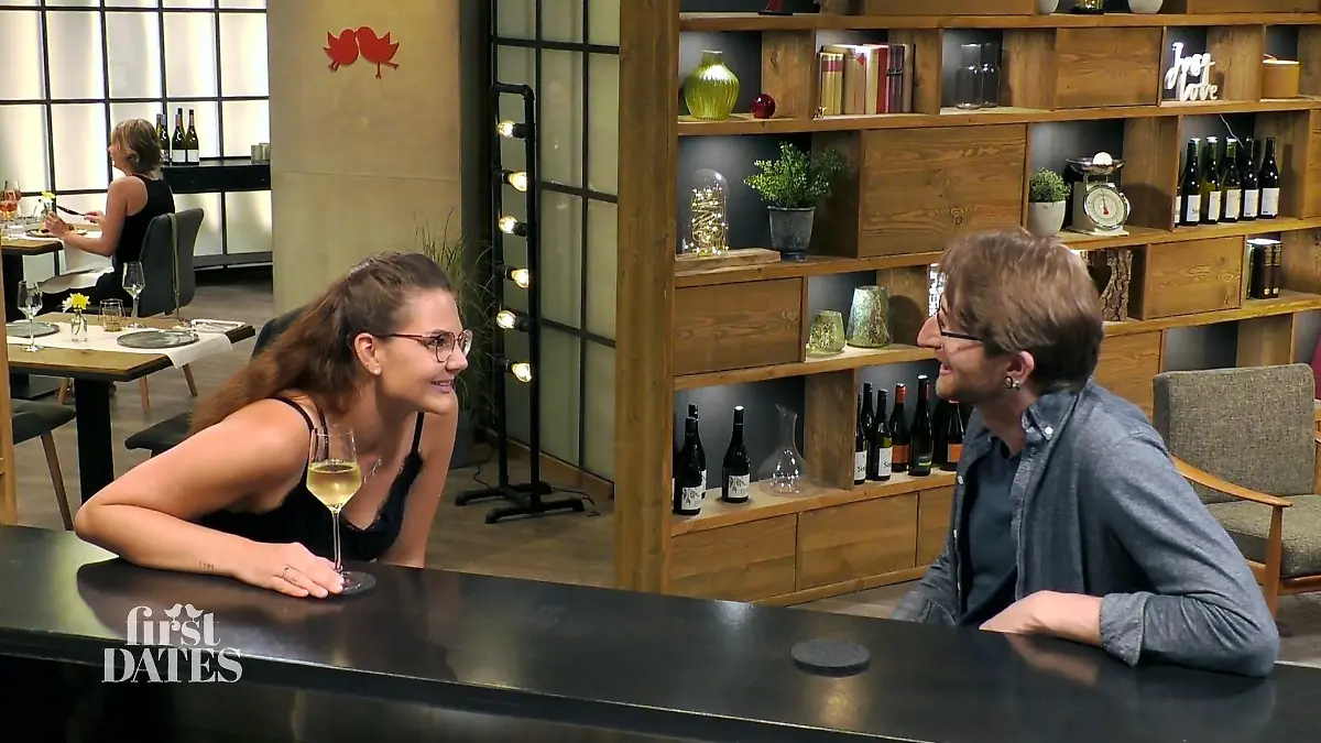 First Dates - Ein Tisch für zwei: Junge Frau mit langen Haaren und Brille und junger Mann mit Brille sitzen sich an der Bar gegenüber und schauen sich interessiert an