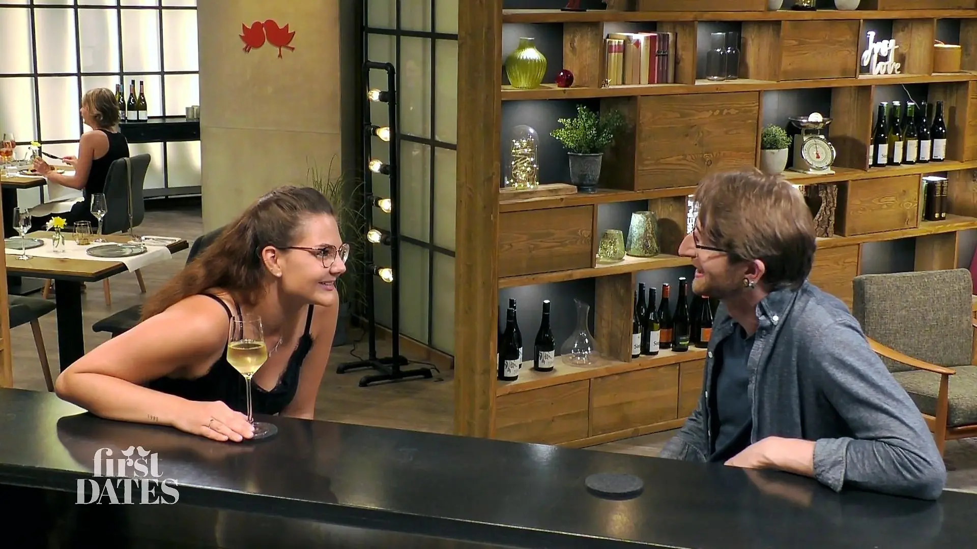 First Dates - Ein Tisch für zwei: Junge Frau mit langen Haaren und Brille und junger Mann mit Brille sitzen sich an der Bar gegenüber und schauen sich interessiert an