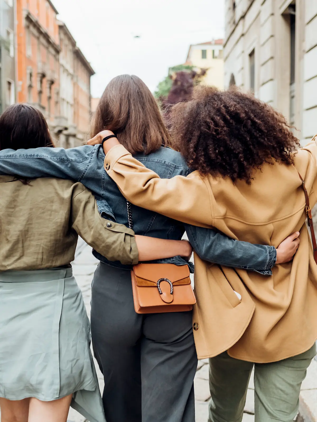Drei Freundinnen sind von hinten zu sehen, wie sie Arm in Arm eine Straße entlanggehen.