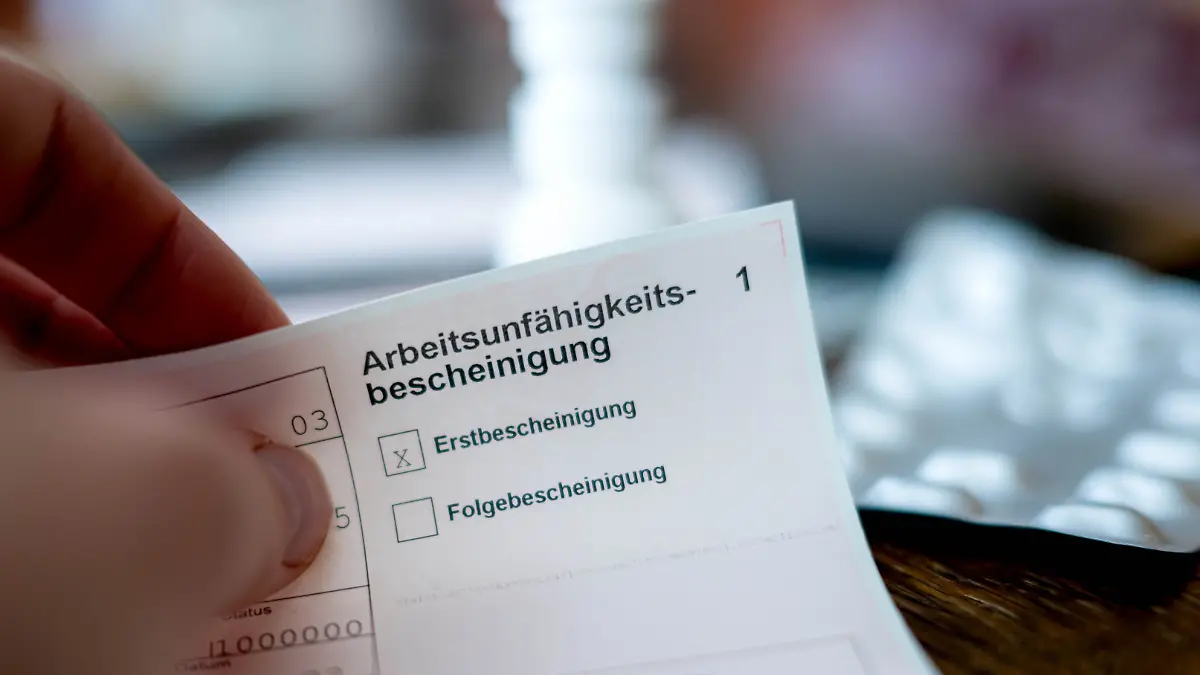Eine Person hält eine ausgefüllte Arbeitsunfähigkeitsbescheinigung in der Hand. Im Hintergrund ist unscharf ein Tisch mit Medikamenten zu sehen. (zu dpa: «Kommission empfiehlt Teilkrankschreibung im Job») +++ dpa-Bildfunk +++