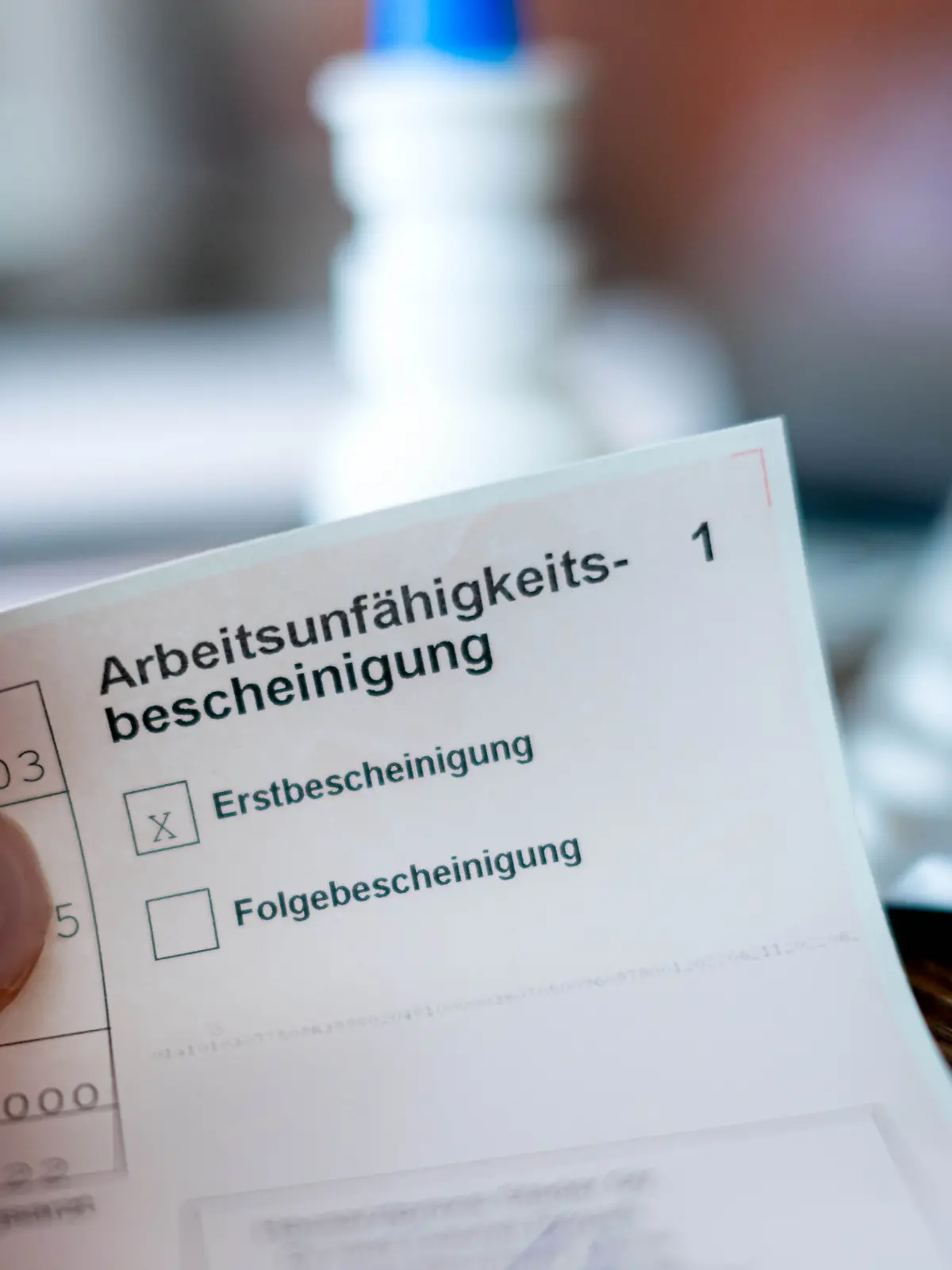 Eine Person hält eine ausgefüllte Arbeitsunfähigkeitsbescheinigung in der Hand. Im Hintergrund ist unscharf ein Tisch mit Medikamenten zu sehen. (zu dpa: «Kommission empfiehlt Teilkrankschreibung im Job») +++ dpa-Bildfunk +++