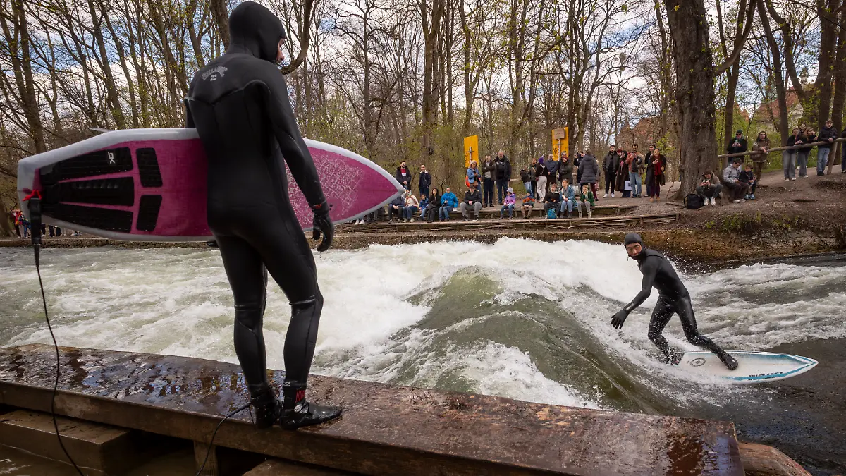 Ein Mann mit Surfbrett schaut einen Surfer zu, der mit seinem Board auf der provisorischen Eisbachwelle im Englischen Garten "reitet". Die Welle verschwand im vergangenen Herbst und hat sich seitdem nicht mehr stabil aufgebaut. Es gab inzwischen mehrere Versuche, sie wieder verlässlich surfbar zu machen. Die Welle ist normalerweise zu jeder Jahres- und Tageszeit ein wahrer Hotspot für Touristen und Surfer aus der ganzen Welt. +++ dpa-Bildfunk +++