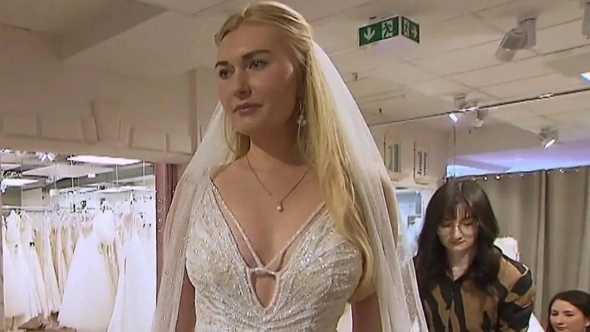 Die Braut betrachtet sich in einem tief ausgeschnittenen Kleid mit Perlenbesatz und Schleier. 