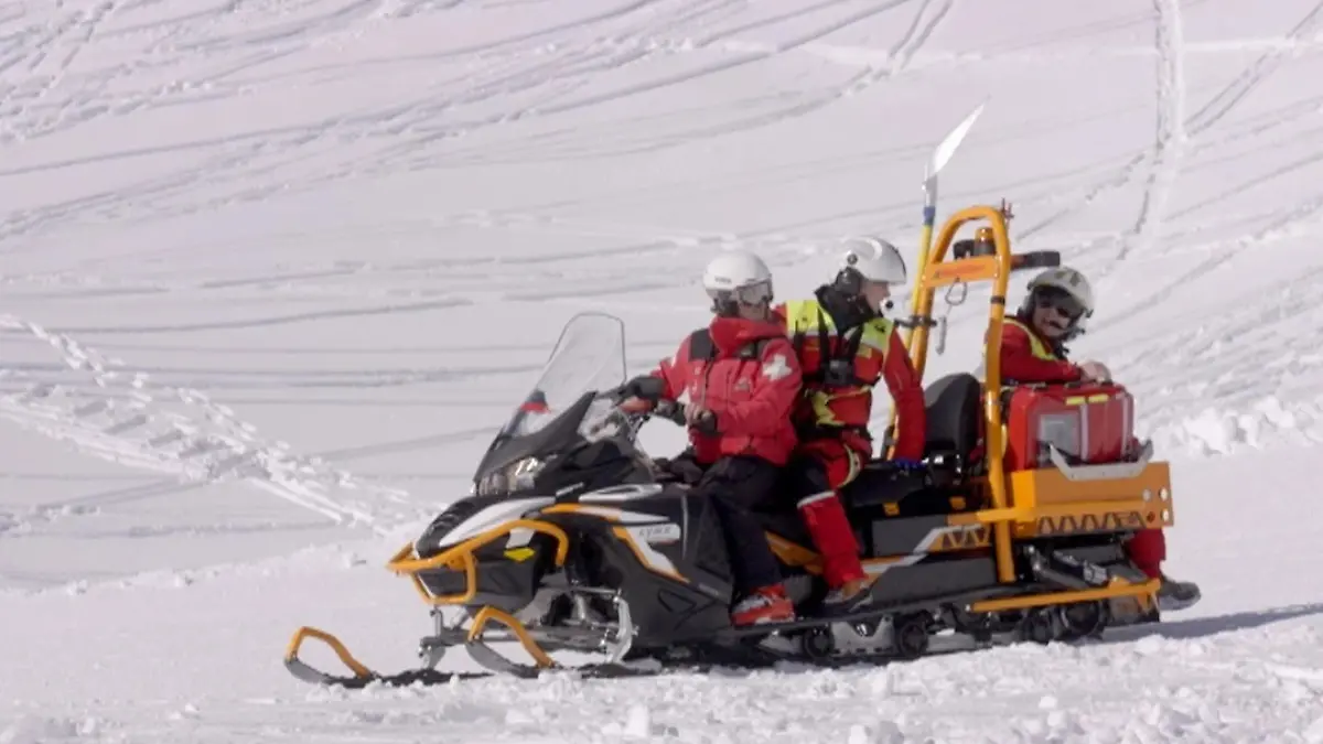 Schneemobil mit drei Rettungskräften auf einer Skipiste.