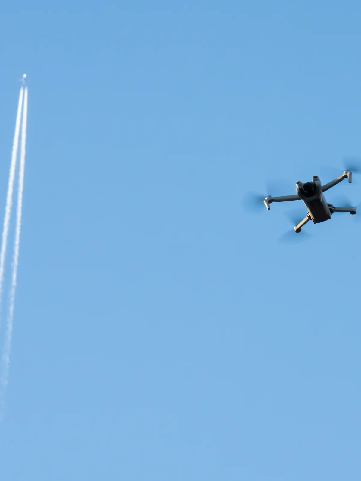 Eine Drohne fliegt im blauen Himmel. Im Hintergrund zieht ein Passagierflugzeug Kondensstreifen hinter sich her.