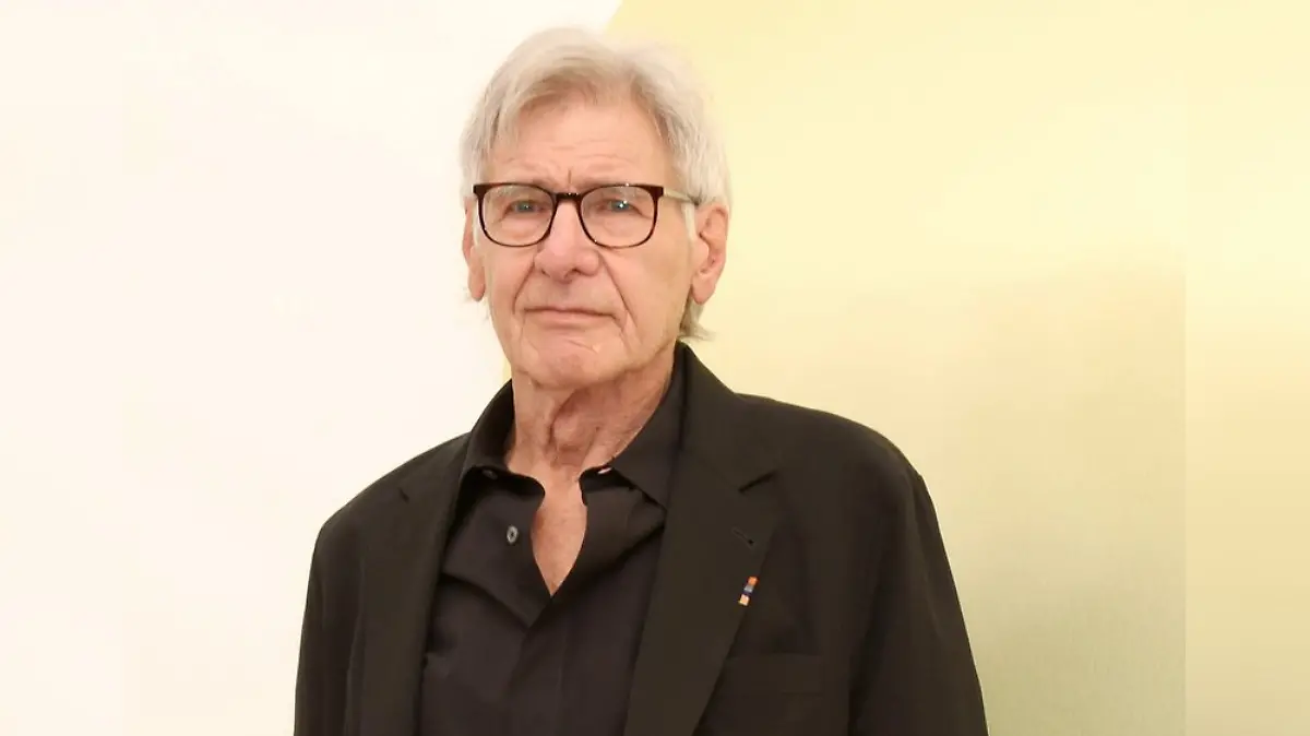 Harrison Ford in einem schwarzen Anzug, mit einer schwarzen Brille, leicht lächelnd, vor einer gelben Wand