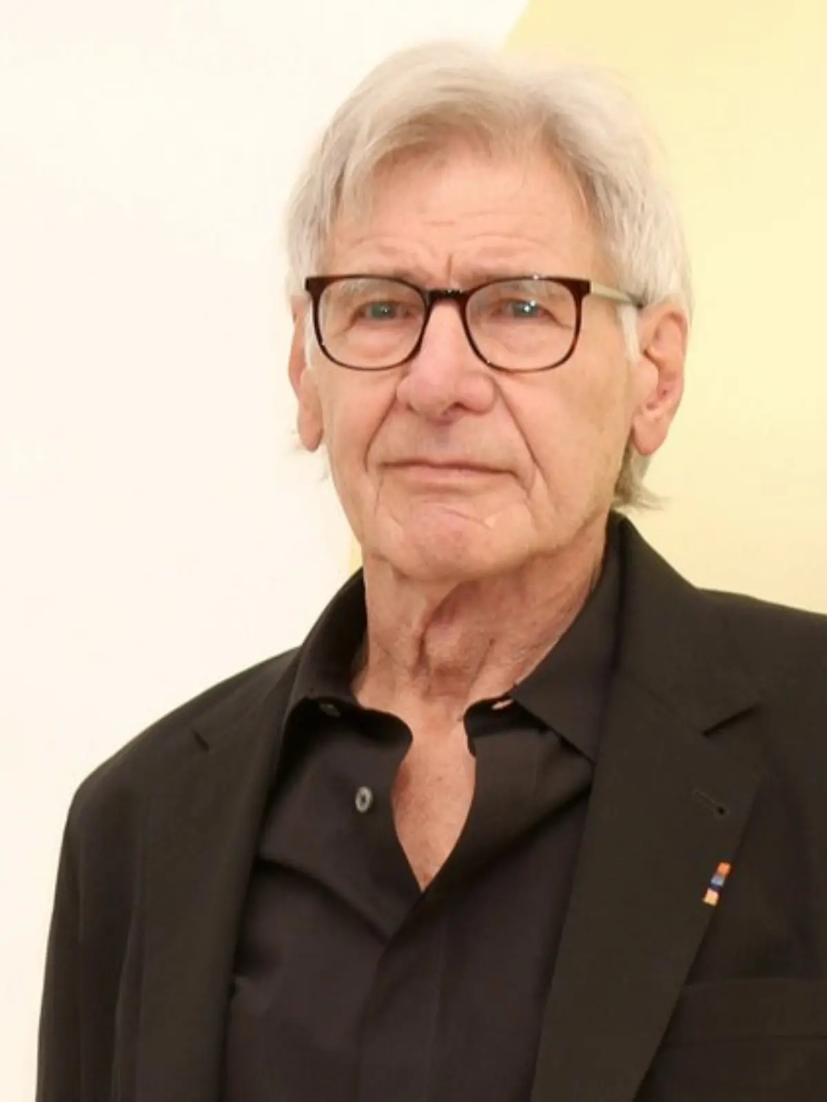 Harrison Ford in einem schwarzen Anzug, mit einer schwarzen Brille, leicht lächelnd, vor einer gelben Wand