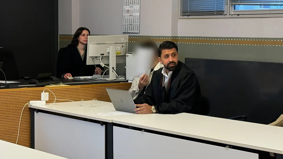 Der Angeklagte sitzt neben seinen Anwalt und hält sich ein Blatt vor das Gesicht.