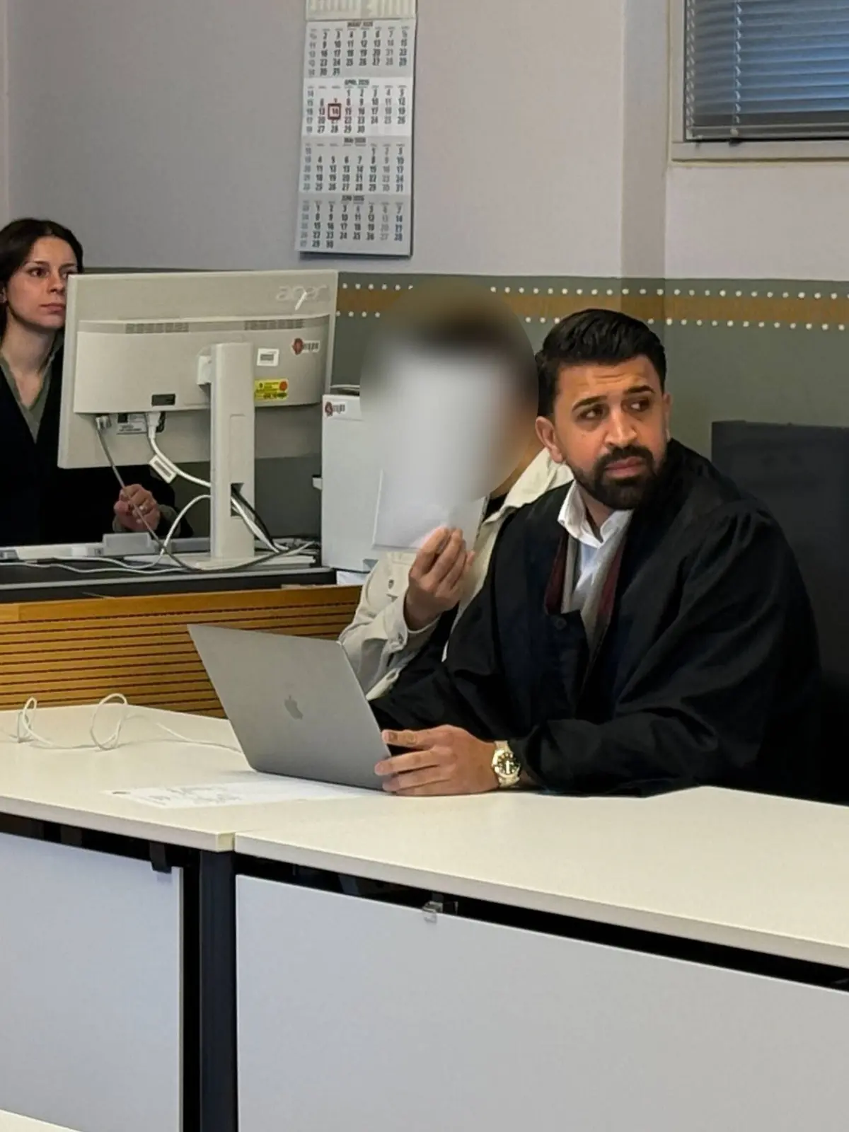 Der Angeklagte sitzt neben seinen Anwalt und hält sich ein Blatt vor das Gesicht.