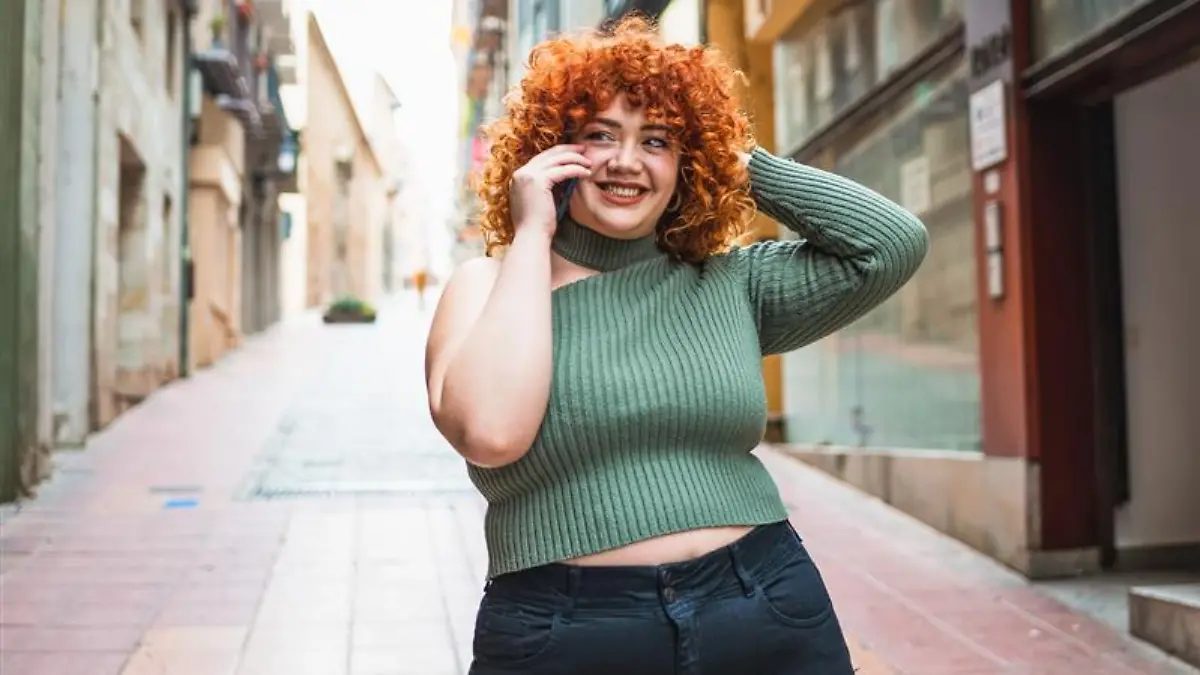 Frau mit lockigen Haaren und frühlingshaftem Outfit steht auf einer Straße und telefoniert.