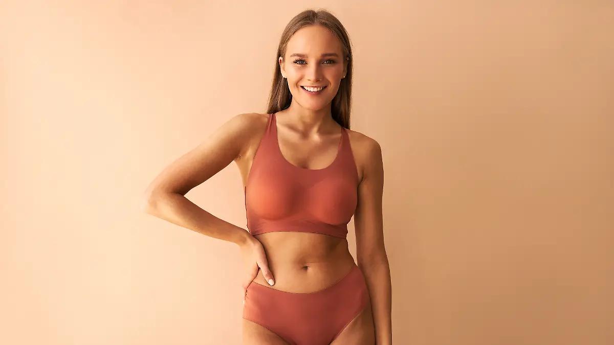 Junge Frau mit gebräunter Haut und blonden Haaren posiert in bequemer Unterwäsche vor einem orangenen Hintergrund, auf ihrem Gesicht ein breites Lächeln