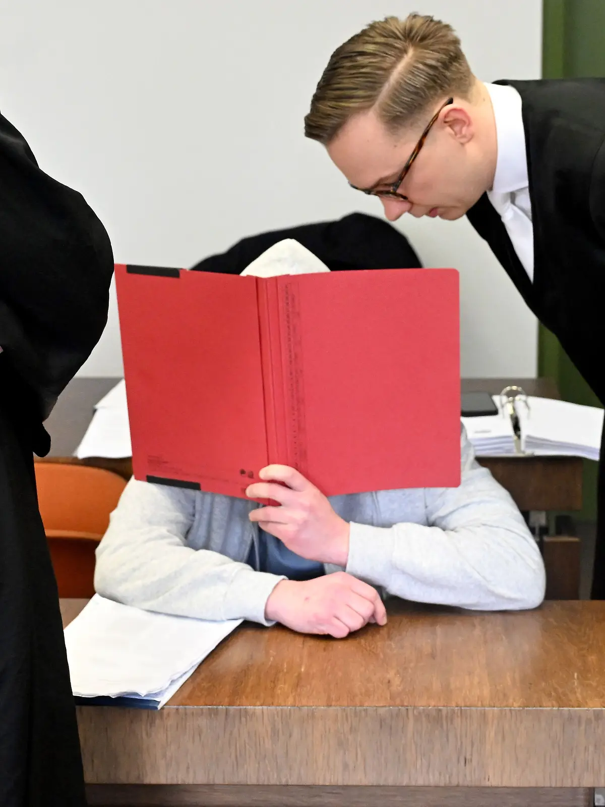 ARCHIV - 18.02.2026, Bayern, München: Der Angeklagte sitzt mit einem Ordner vor dem Gesicht vor Beginn des Prozess wegen Mordes an seiner Großmutter im Gerichtssaal im Landgericht München. Der Mann soll vor knapp einem Jahr seine Großmutter aus Habgier getötet haben. Die Großmutter hatte ihn stets unterstützt, auch finanziell. Als sie mit Kontaktabbruch drohte, soll er sie getötet haben. (zu dpa: «Enkel wegen Mordes an seiner Großmutter verurteilt»)