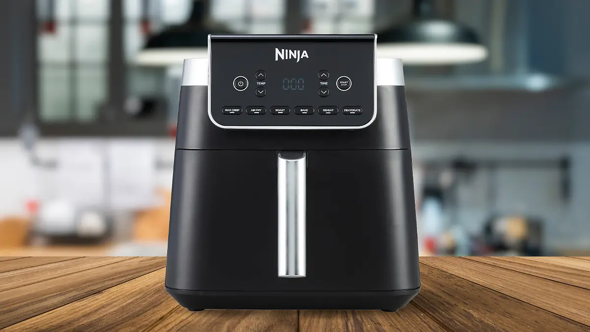 Die Ninja Heißluftfritteuse Max Pro AF180EU auf einem Holztisch, die Küche im Hintergrund ist verschwommen.