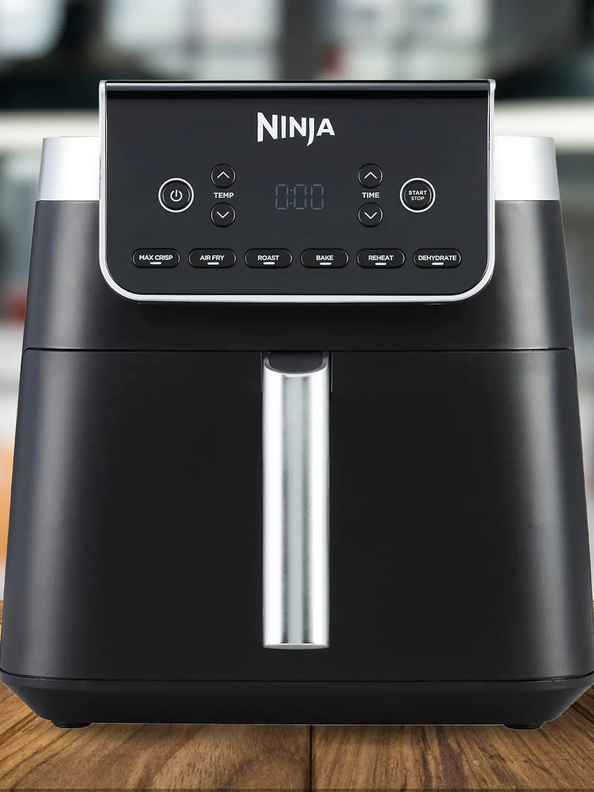 Die Ninja Heißluftfritteuse Max Pro AF180EU auf einem Holztisch, die Küche im Hintergrund ist verschwommen.