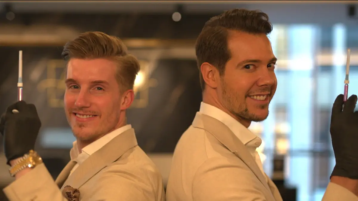 Für immer jung: Zwei Männer in hellen Anzügen und Spritze in der Hand lächeln Rücken an Rücken in die Kamera