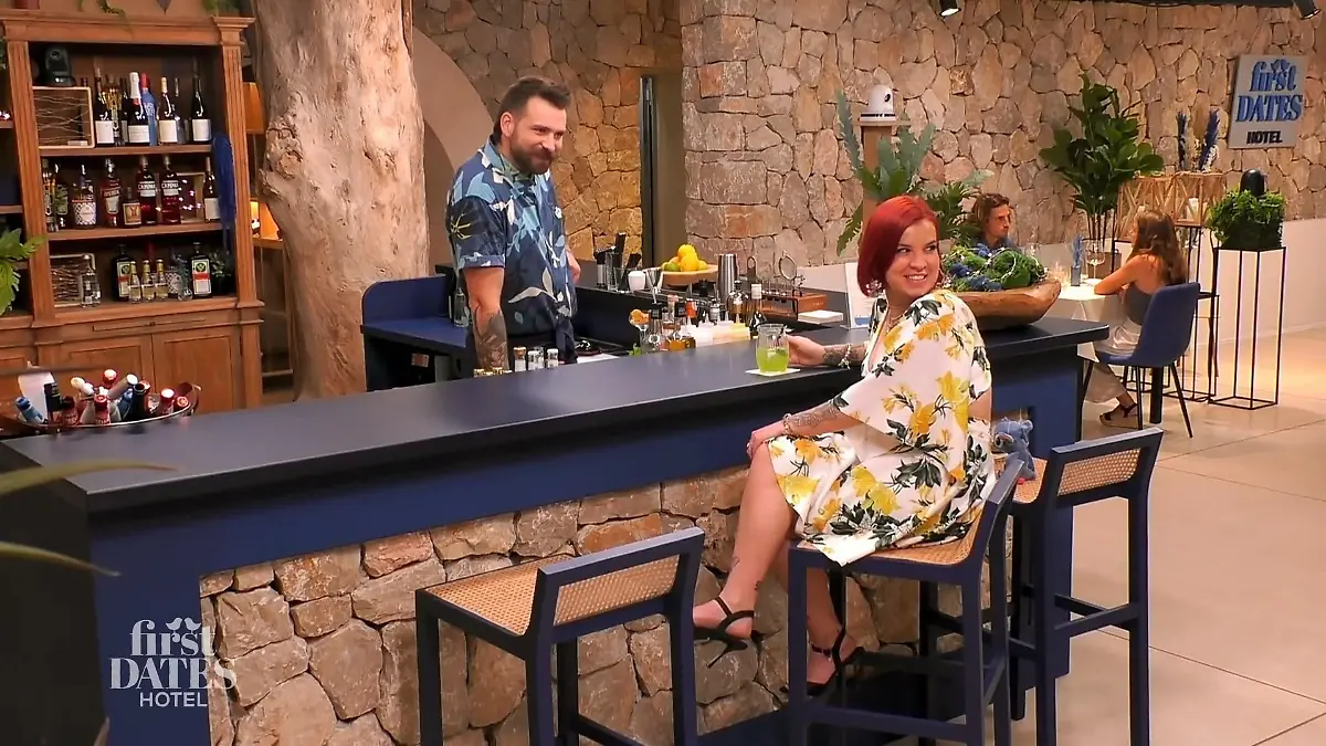 Denise sitzt an der Bar und schaut über die Schulter.