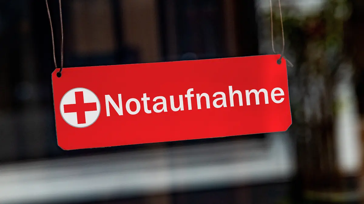 [M] Ein symbolisches, rotes Schild mit einem roten Kreuz sowie der Aufschrift "Notaufnahme" haengt leicht schraeg an Lederbaendern in einem Fenster. Foto mit Composing als Symbolbild fuer die Notfallversorgung in Krankenhaeusern. Oft werden die Notaufnahmen auch von Leuten zur Behandlung aufgesucht, die keine Notfaelle sind. Die Notaufnahme ist fuer Patienten mit Lebensbedrohlichen Zustaende, schweren Verletzungen oder akuten, schweren Erkrankungen.