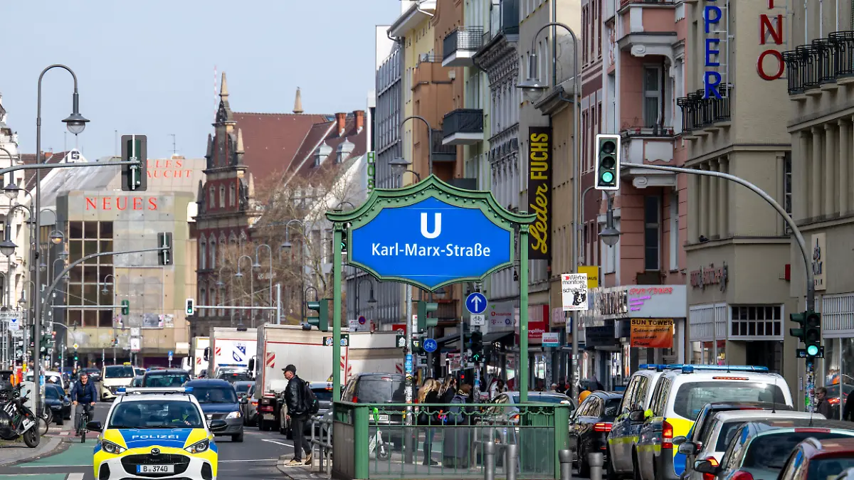 Das Eingangsschild zum U-Bahnhof Karl-Marx-Straße steht an der gleichnamigen Straße im Berliner Stadtteil Neukölln, während Fahrzeuge im Straßenverkehr an den Eingängen zur U-Bahn vorbeifahren.