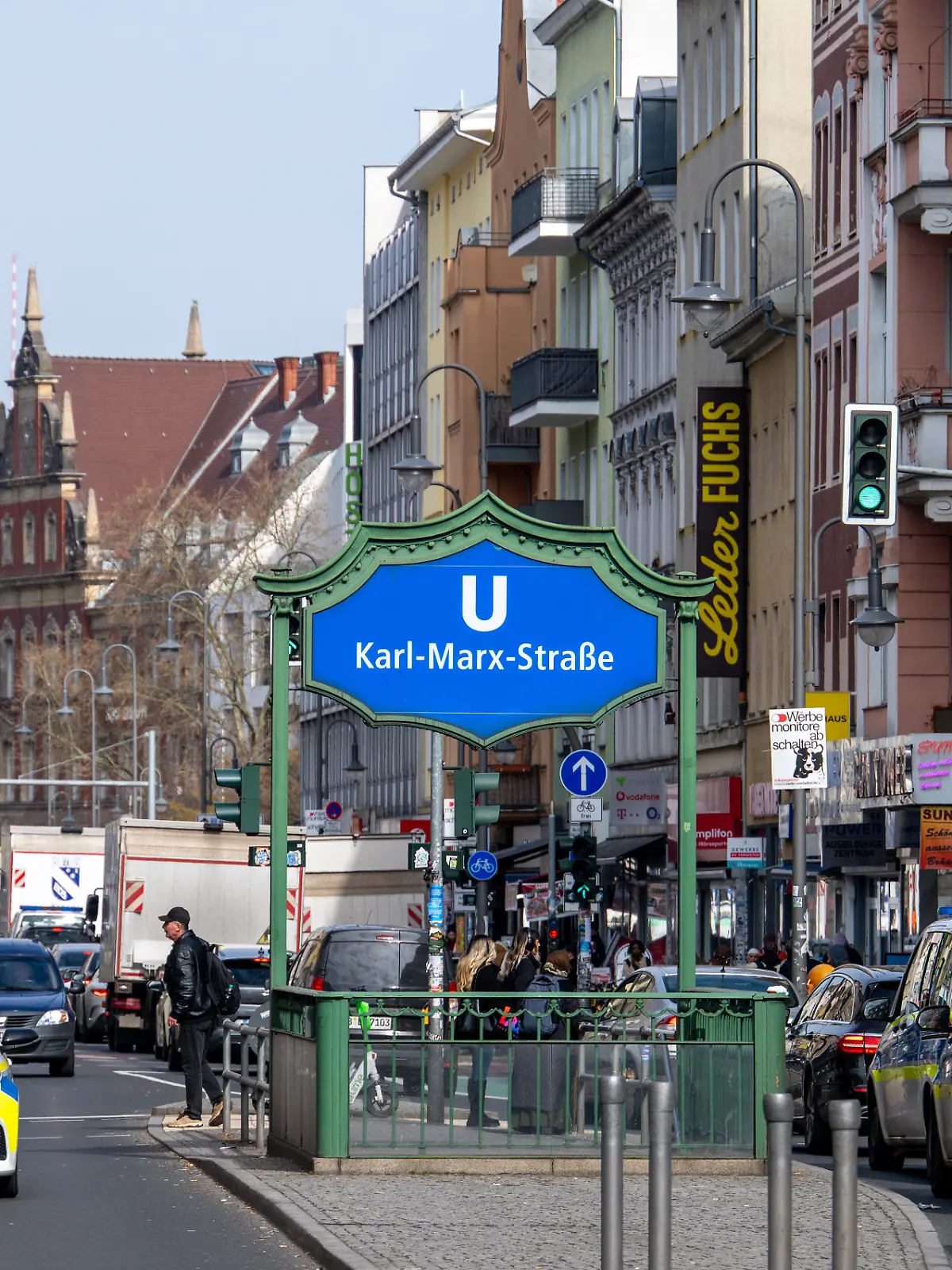 Das Eingangsschild zum U-Bahnhof Karl-Marx-Straße steht an der gleichnamigen Straße im Berliner Stadtteil Neukölln, während Fahrzeuge im Straßenverkehr an den Eingängen zur U-Bahn vorbeifahren.