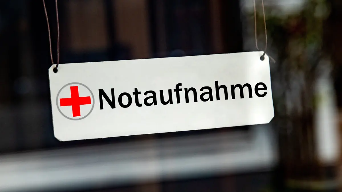 [M] Ein symbolisches, silbernes / weisses Schild mit einem roten Kreuz sowie der Aufschrift "Notaufnahme" haengt leicht schraeg an Lederbaendern in einem Fenster. Foto mit Composing als Symbolbild fuer die Notfallversorgung in Krankenhaeusern. Oft werden die Notaufnahmen auch von Leuten zur Behandlung aufgesucht, die keine Notfaelle sind. Die Notaufnahme ist fuer Patienten mit Lebensbedrohlichen Zustaende, schweren Verletzungen oder akuten, schweren Erkrankungen.