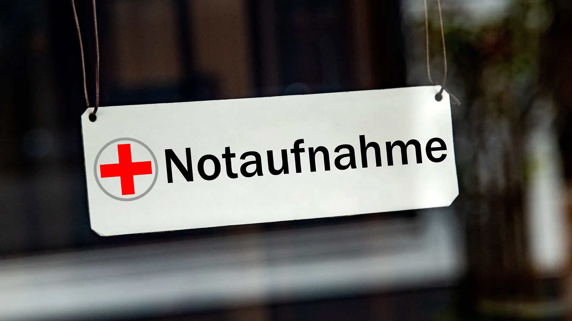 [M] Ein symbolisches, silbernes / weisses Schild mit einem roten Kreuz sowie der Aufschrift "Notaufnahme" haengt leicht schraeg an Lederbaendern in einem Fenster. Foto mit Composing als Symbolbild fuer die Notfallversorgung in Krankenhaeusern. Oft werden die Notaufnahmen auch von Leuten zur Behandlung aufgesucht, die keine Notfaelle sind. Die Notaufnahme ist fuer Patienten mit Lebensbedrohlichen Zustaende, schweren Verletzungen oder akuten, schweren Erkrankungen.