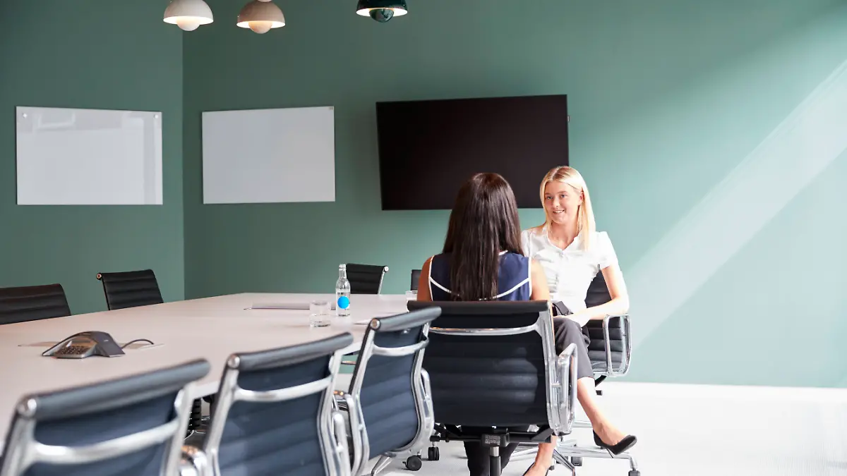 Bewerbungsgespräch unter vier Augen, zwei Frauen sitzen sich lächelnd gegebenüber in einem Büro mit einem langen Tisch und vielen Stühlen