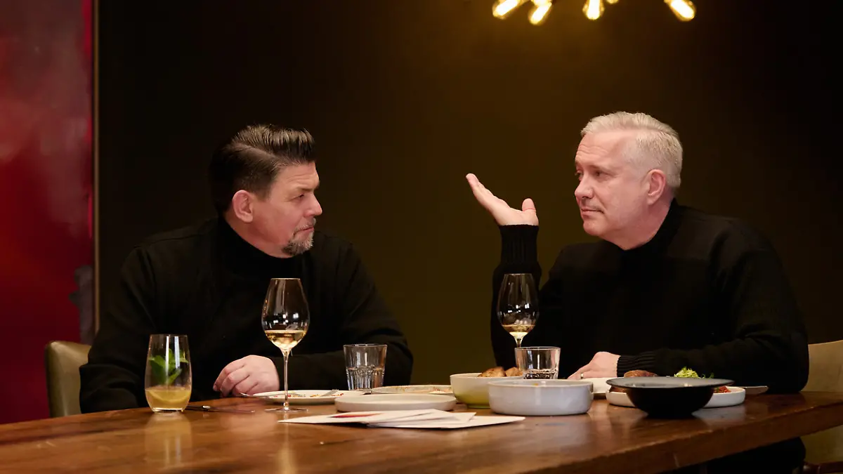 Tim Mälzer und Alexander Herrmann in einem dunklen Raum am Tisch sitzend. 