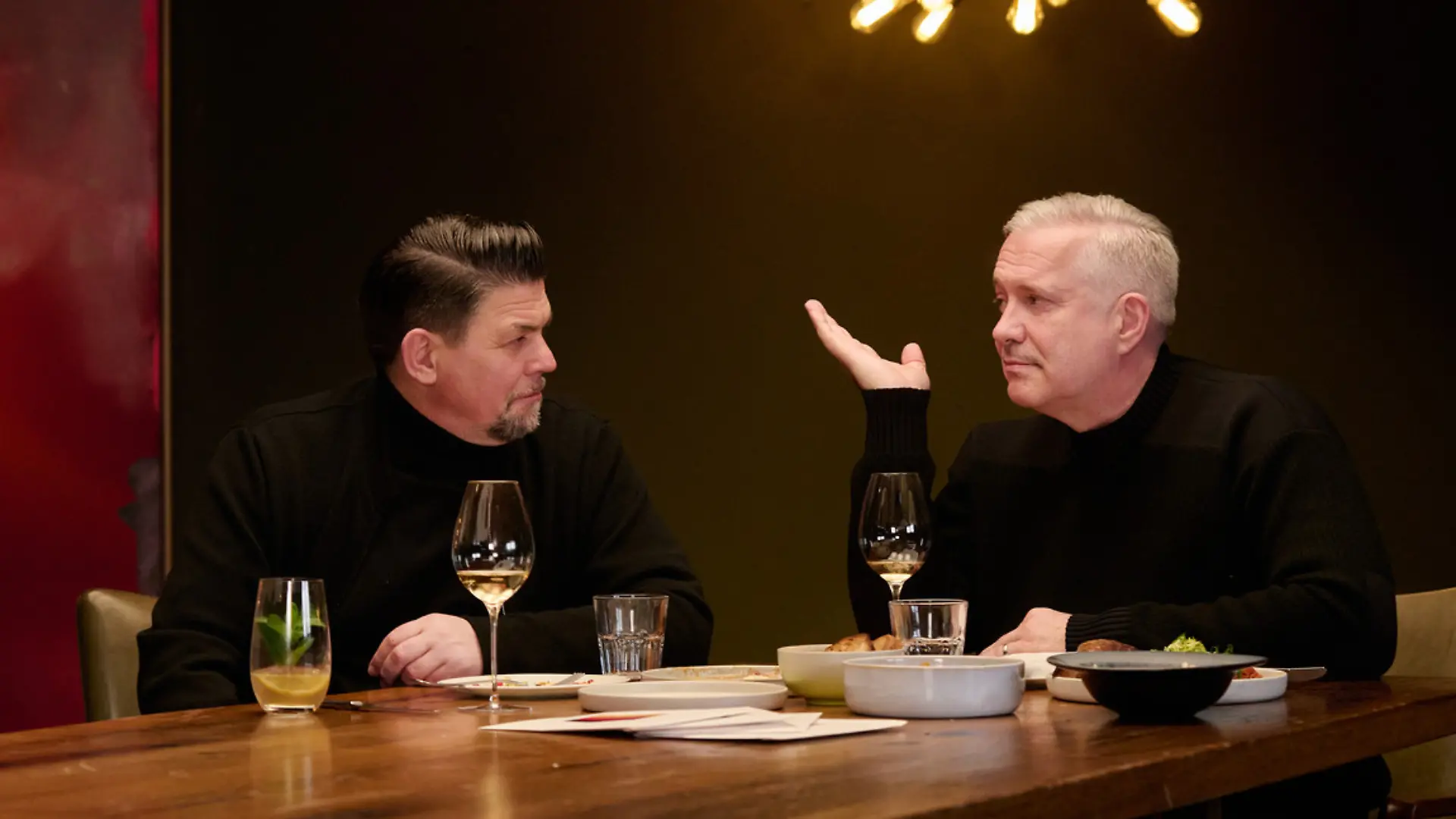 Tim Mälzer und Alexander Herrmann in einem dunklen Raum am Tisch sitzend.